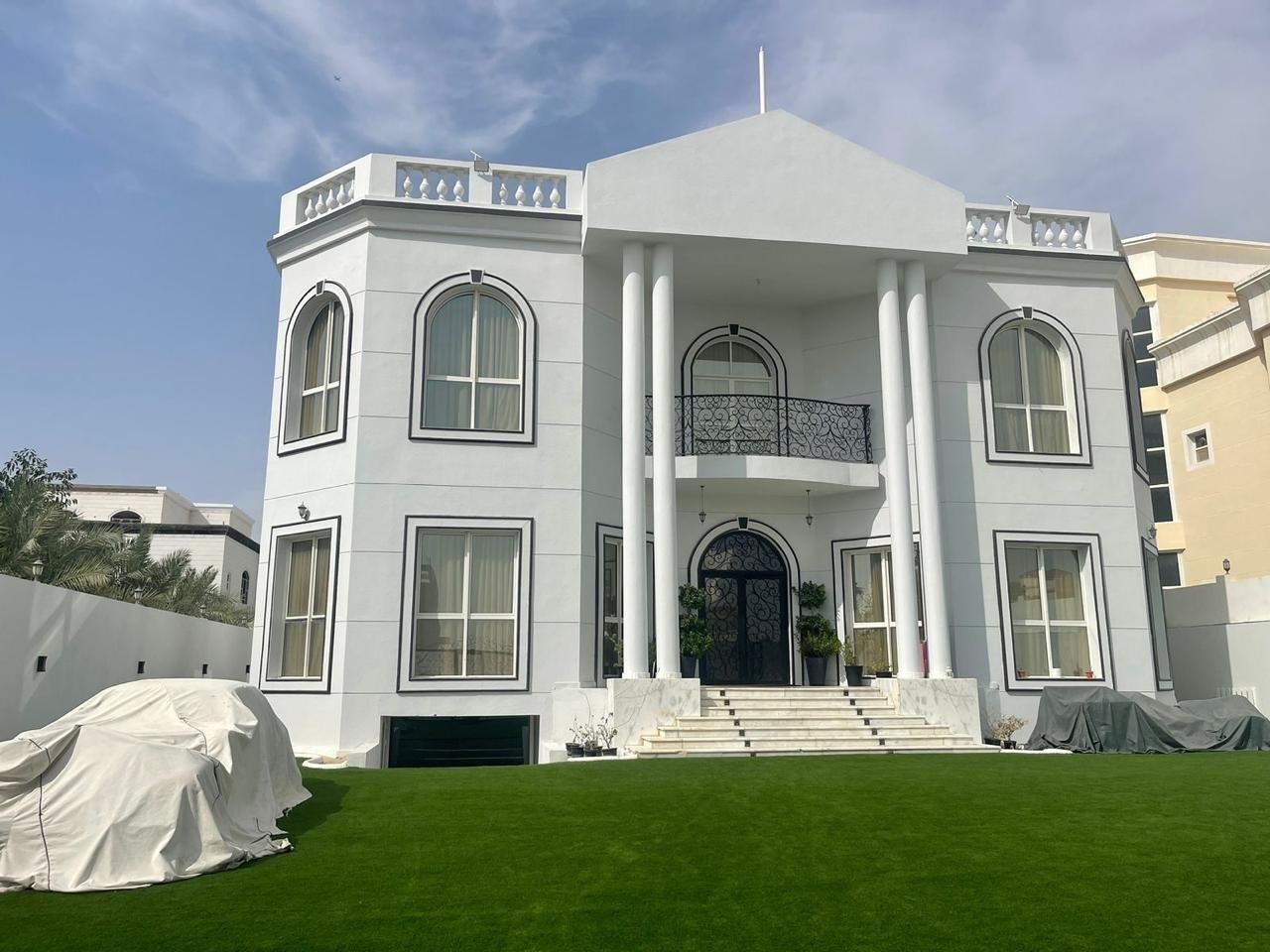 White neo-classical villa facade with columned portico, seen across a walled lawn in daylight.